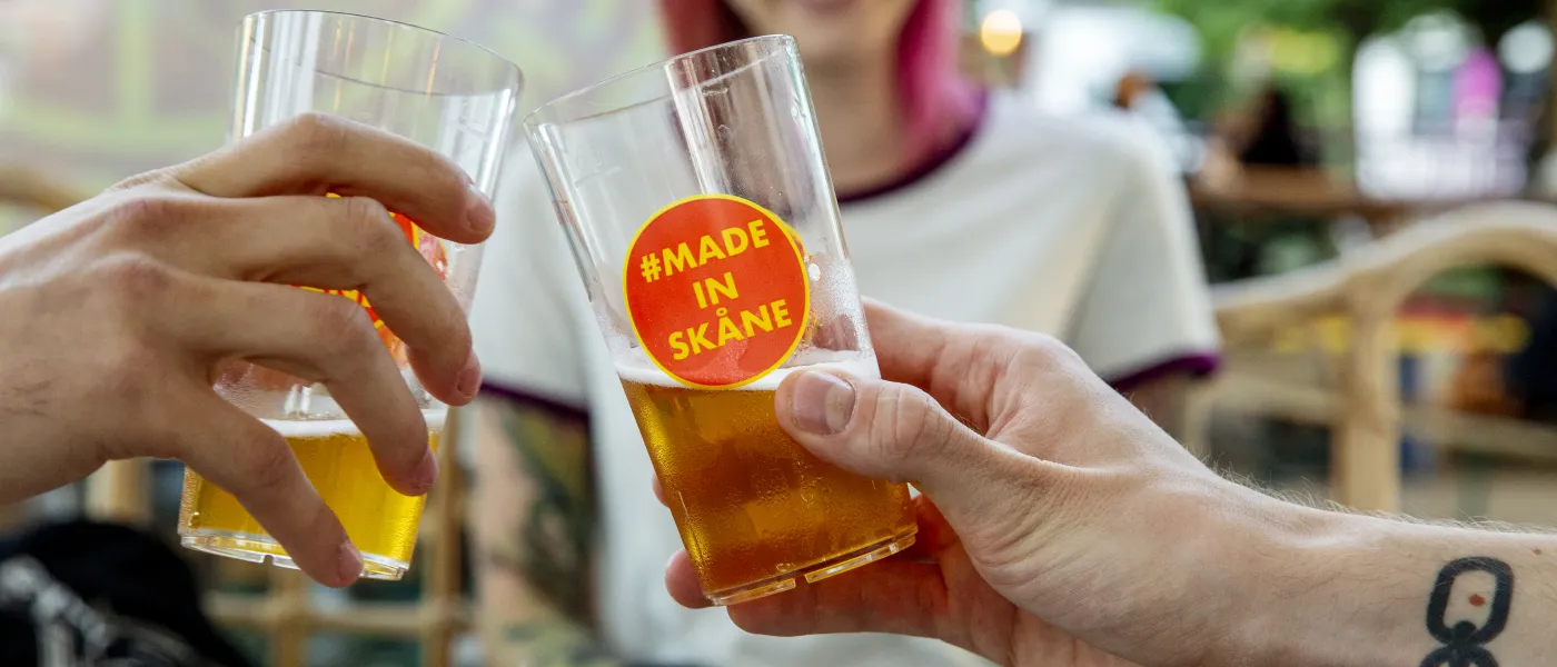 Två händer som håller i ölglas med texten made in skåne möts i en skål. En leende person står i bakgrunden
