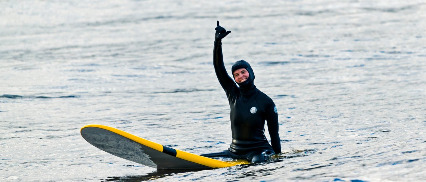 Kvinna i torrdräkt sitter på gul surfbräda med handen upp i hälsning