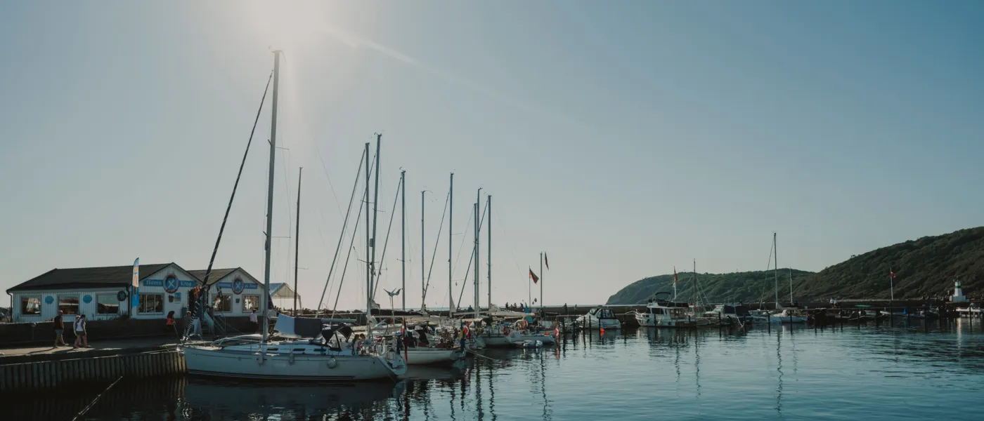 Solglittrande hamn med segelbåtar vid kaj, klarblå himmel och grönskande kulle i bakgrunden