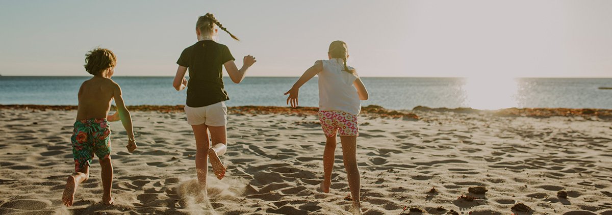 Tre barn springer mot havet på en strand. Foto.