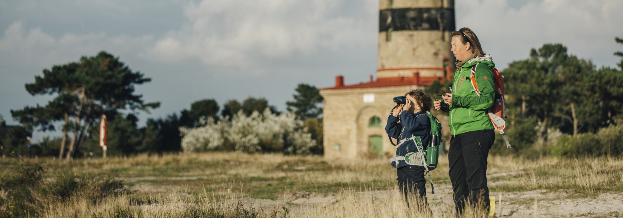 En vuxen person och ett barn med kikare står på en strand med en fyr i bakgrunden