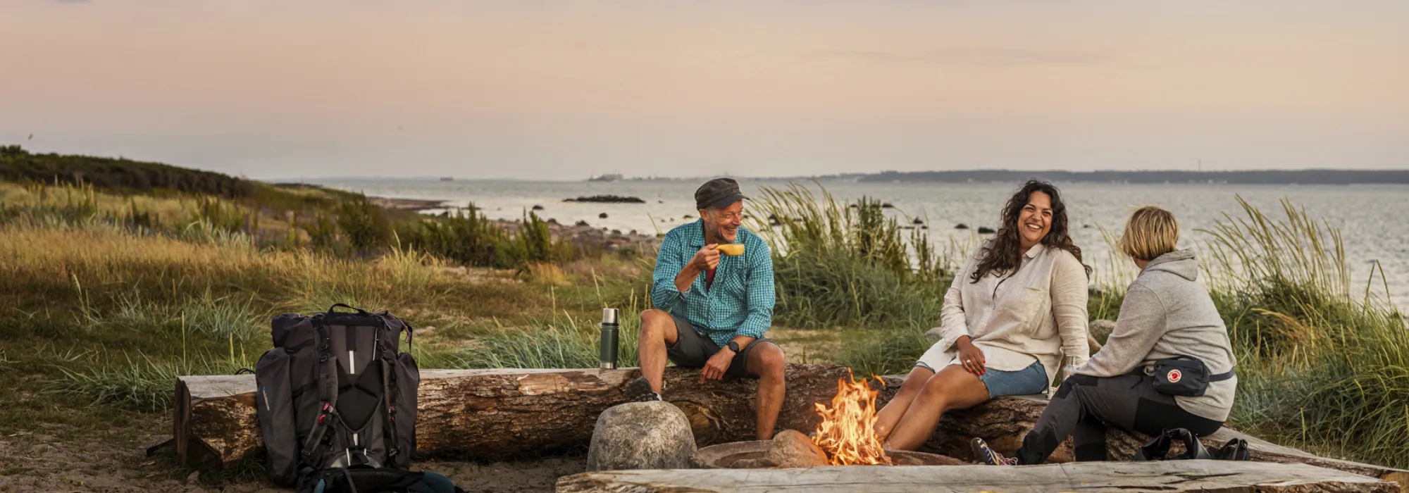 En man och två kvinnor sitter vid en eldstad vid havet