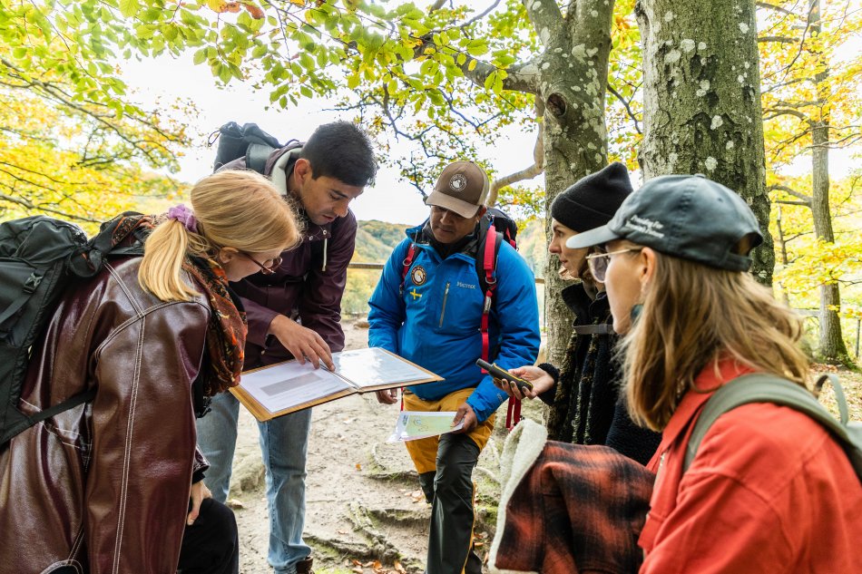 Fem av deltagarna i uppdrag fotspår på plats i Söderåsens nationalpark. De står böjda över en karta och söker efter rätt väg. Foto.