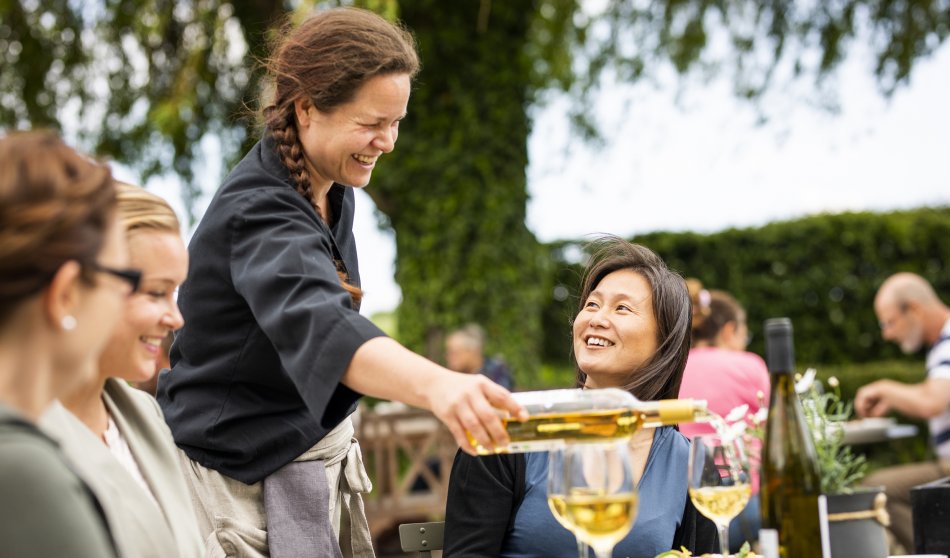 En kvinna serverar vin till ett sällskap som sitter vid ett bord utomhus. Foto.