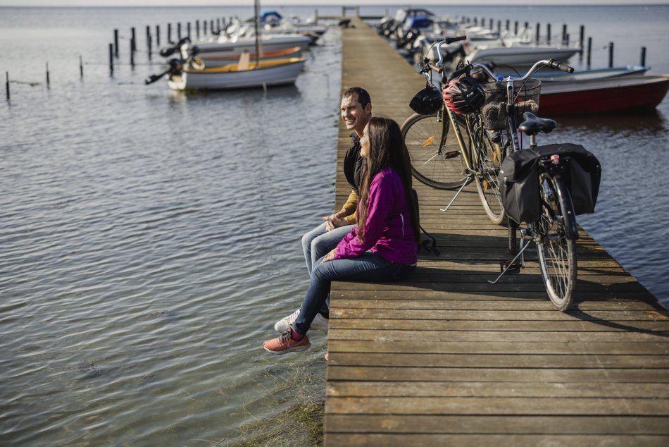 Två personer sitter på en brygga och dinglar med benen över vatten. Bakom sig har de sina cyklar parkerade. Foto.