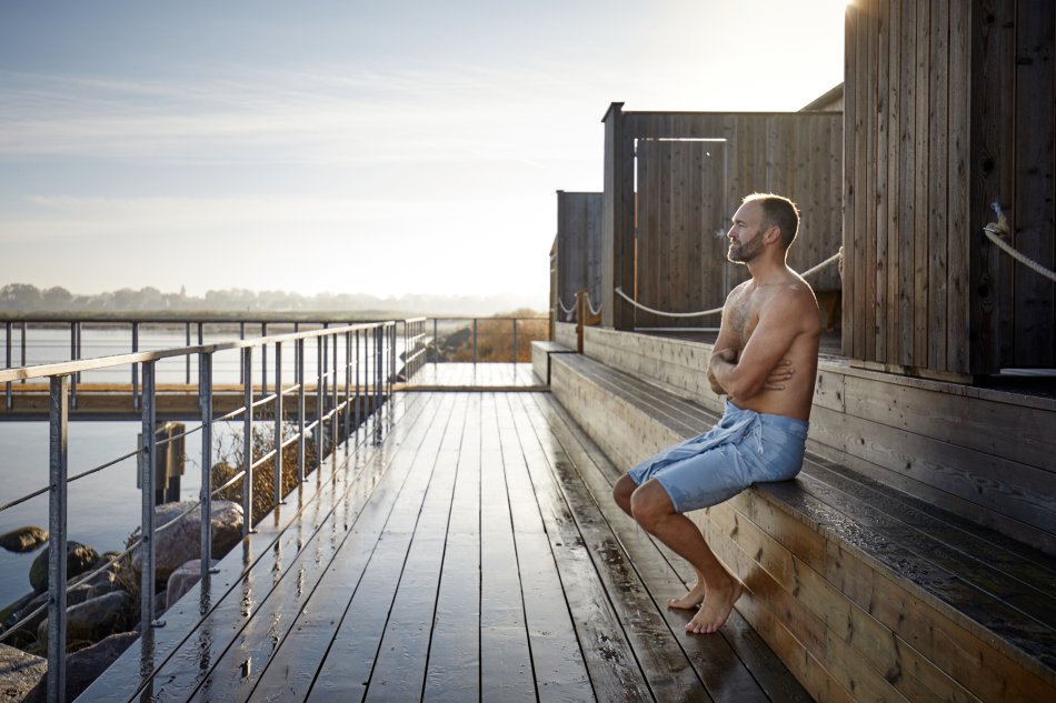En man sitter på bryggan vid ett kallbadhus. 
