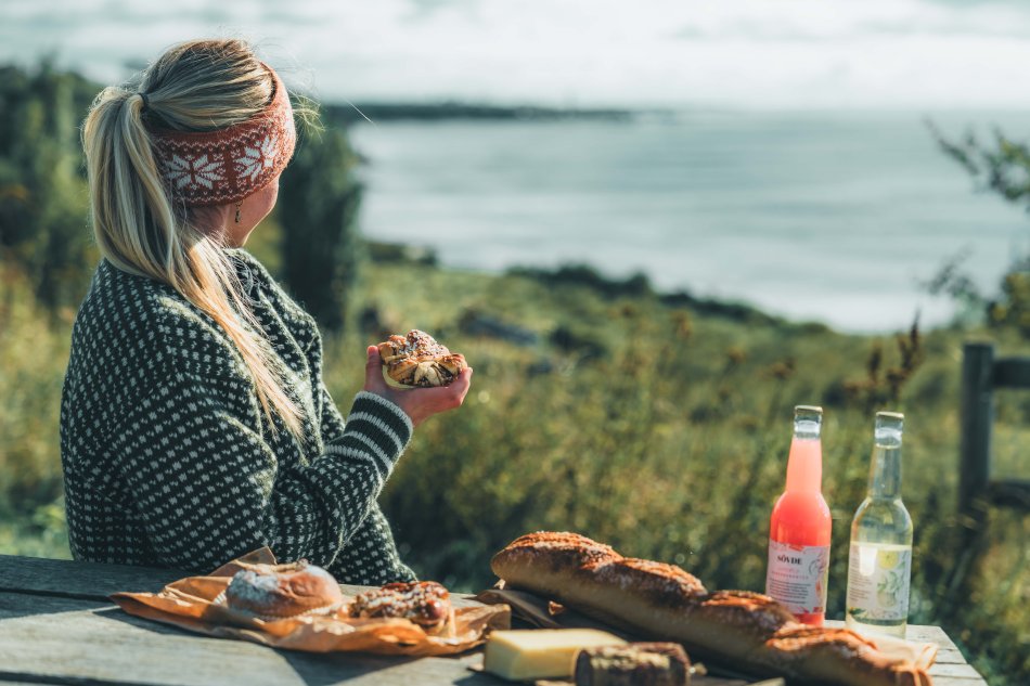 Kvinna i pannband och stickad grov tröja blickar ut över havet och fikar på bullar och dricka